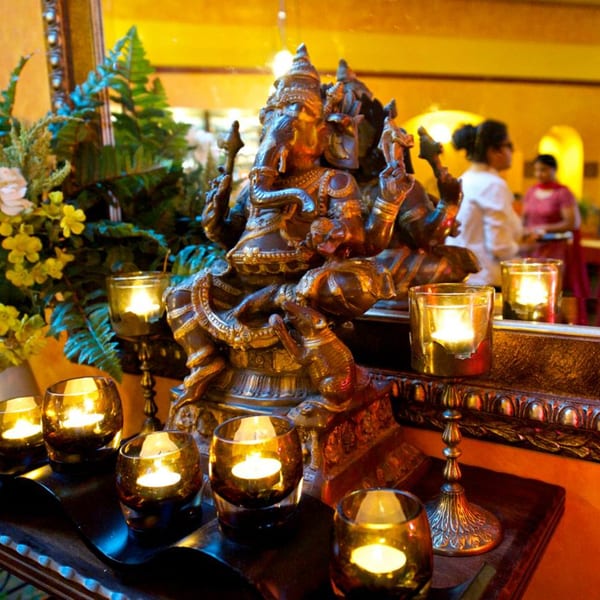 Bronze Ganesha statue surrounded by lit votive candles and greenery, with blurred people in the background.