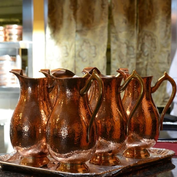 Five hammered copper water pitchers with brass handles on a silver tray at Delhi Diner.