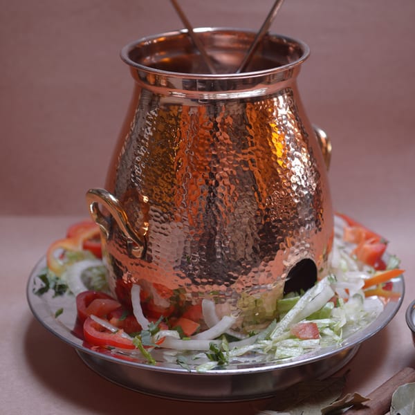 A hammered copper Indian serving pot with two spoons, surrounded by a fresh salad on a silver platter.