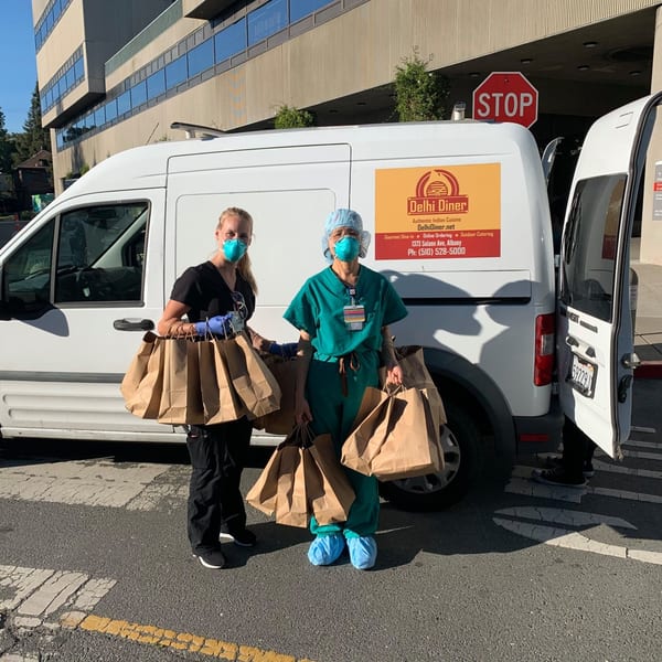 Two masked healthcare workers hold Delhi Diner food bags next to a delivery van outside a building.