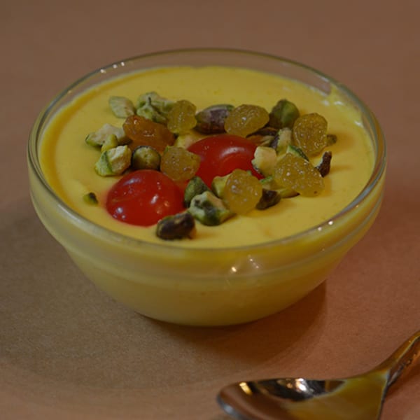 A glass bowl of yellow Indian dessert, topped with pistachios, raisins, and red cherries.