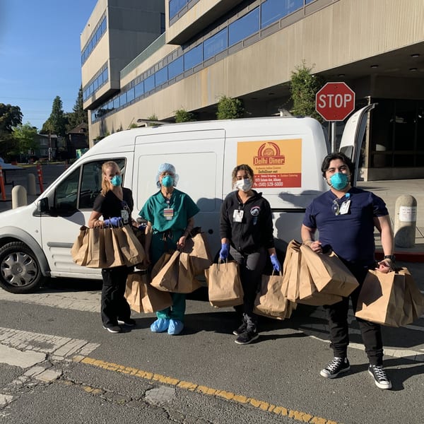 Four masked Delhi Diner staff members hold many Indian food takeout bags in front of a delivery van.