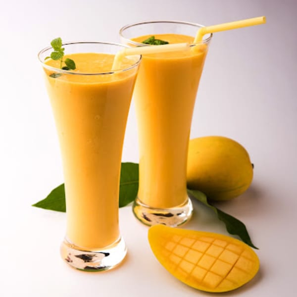 Two glasses of mango lassi with mint, whole mango, and sliced mango on a white surface.