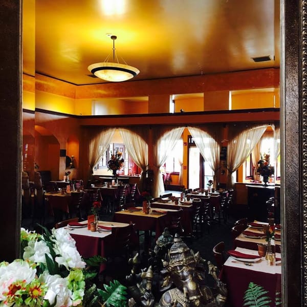 Warmly lit Indian restaurant interior with set tables, arched windows, and decorative statues.